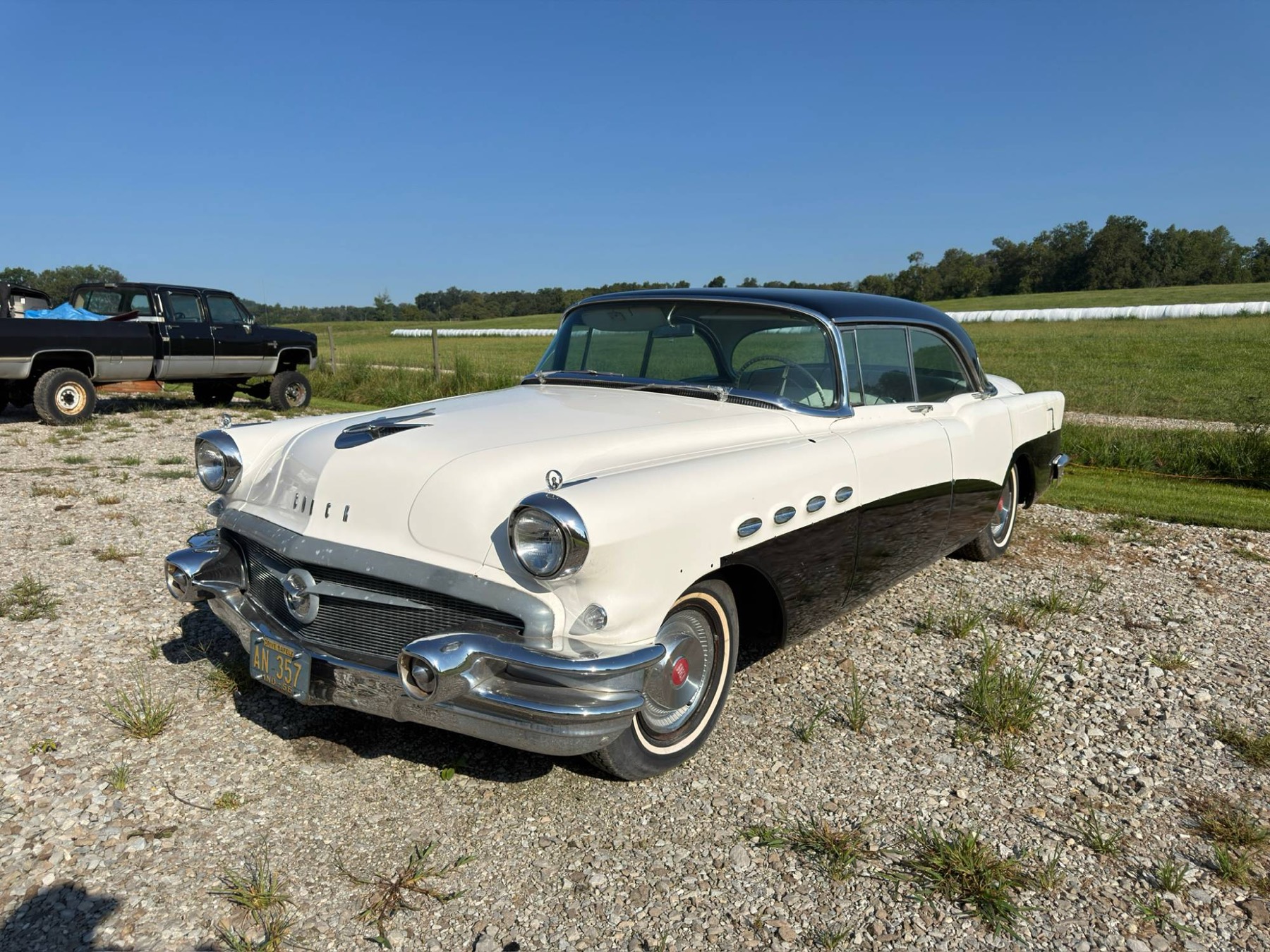 White and black 1956 Buick parked on gravel in front of a green field